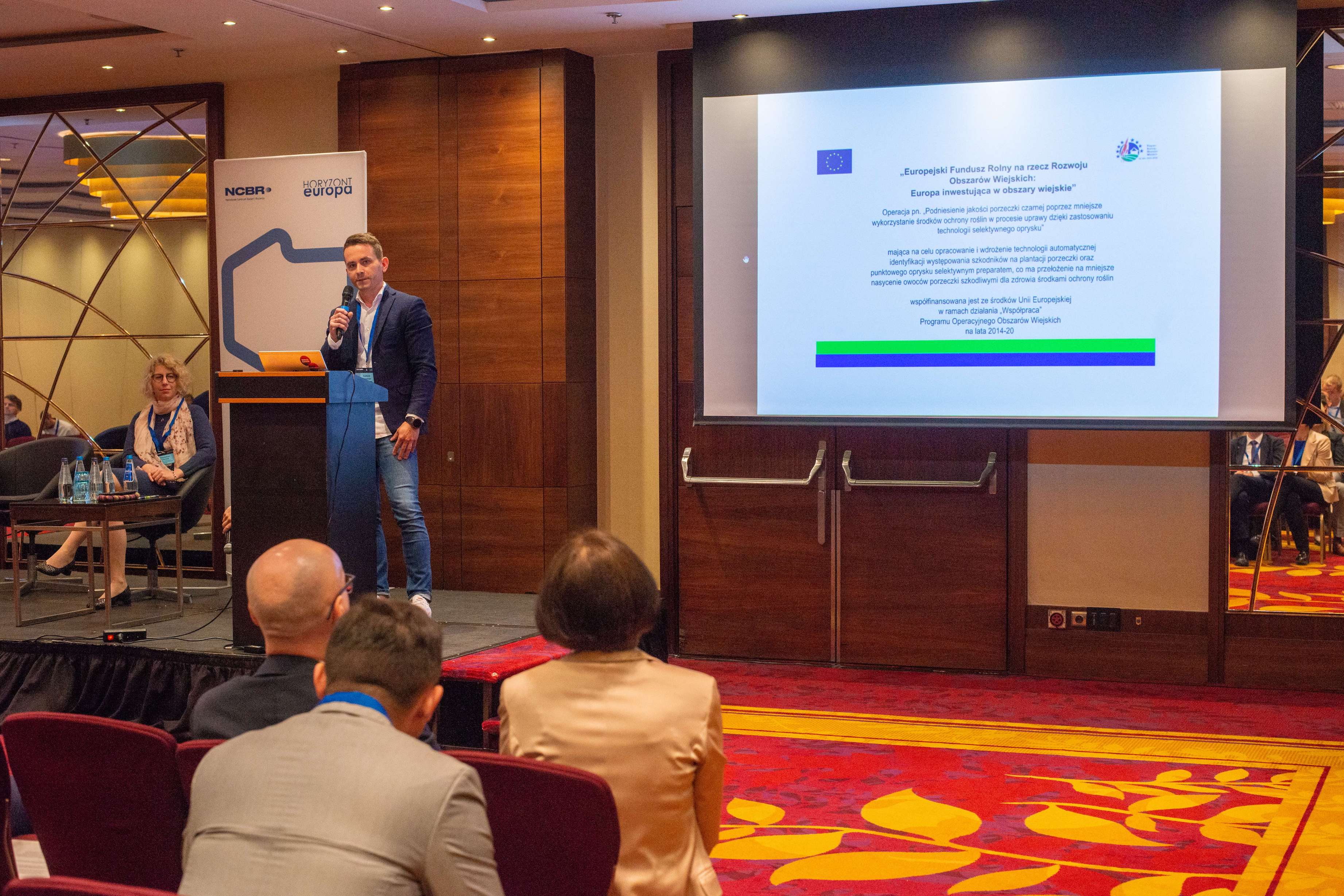 CEO Łukasz Kopiński prezentuje projekt 'Owoce 4.0' na konferencji organizowanej przez NCBiR. W tle prezentacja ze slajdem od dofinansowaniu pochodzącym z ARiMRu.
